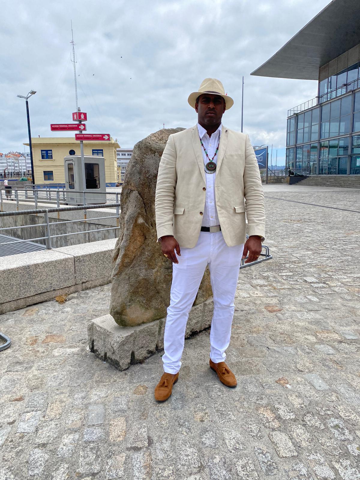 Awo Ifalade con sombrero y traje claro, en un paseo urbano junto al puerto.