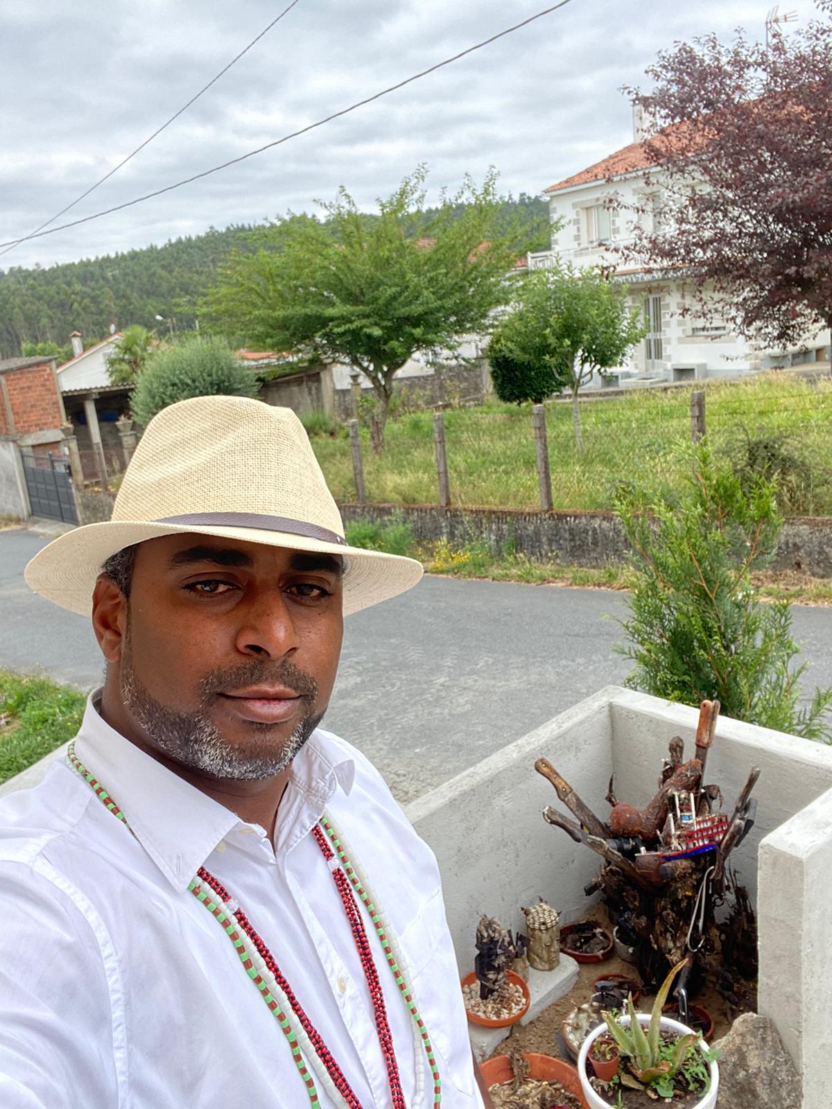 Awo Ifalade con sombrero y collares, junto a elementos de su tradición en un entorno exterior.